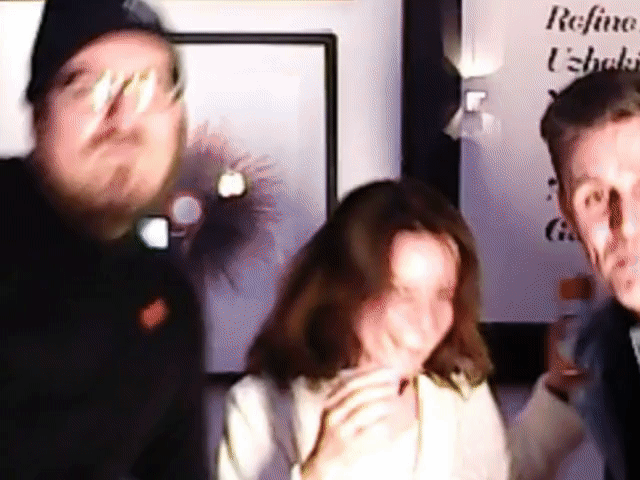 Three blurred people indoors, one holding a drink, with wall art and partial text in the background