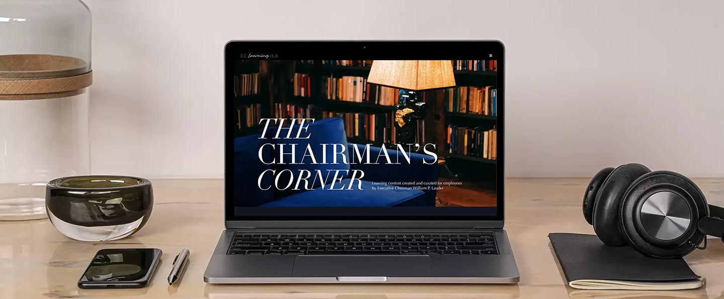 Laptop on desk showing The Chairman's Corner, with phone, pen, headphones, notebook, and glassware