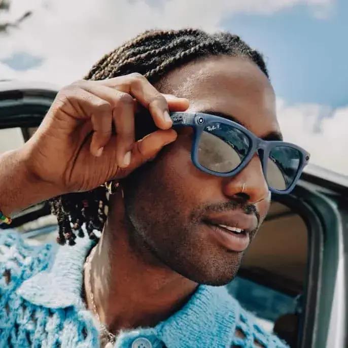 Man adjusting blue sunglasses while leaning out of a car under a bright sky