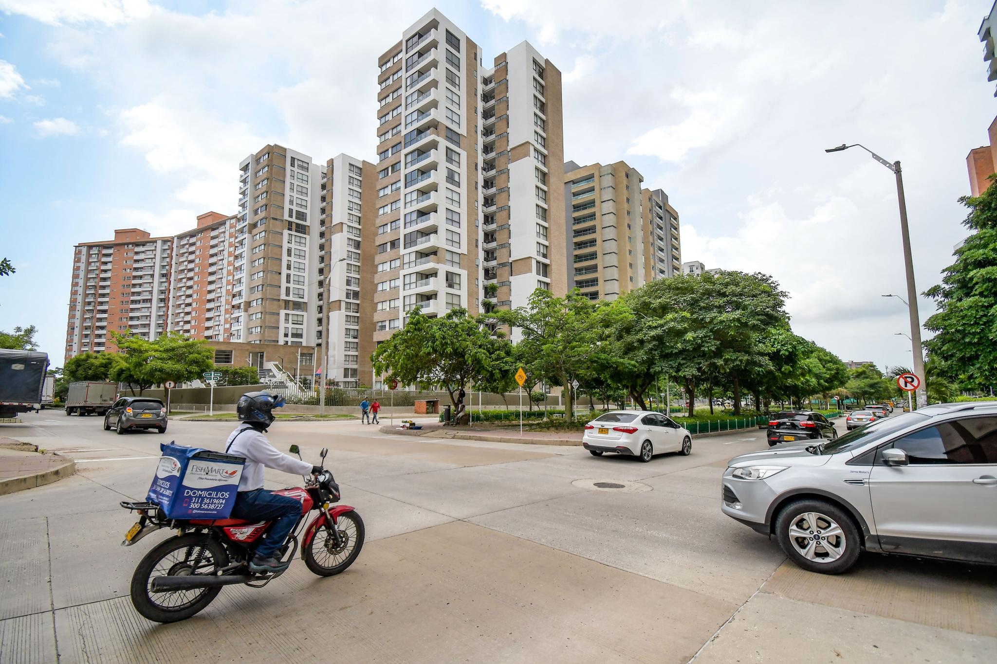 Urban intersection with high-rise apartments, cars, trees, and a delivery motorcyclist
