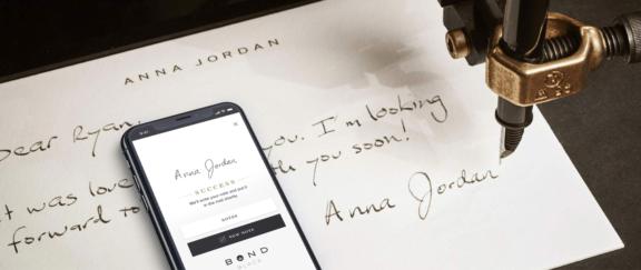 Bond Black D Cover Robotic pen handwriting a personal note while a phone shows the Bond Black app confirmation