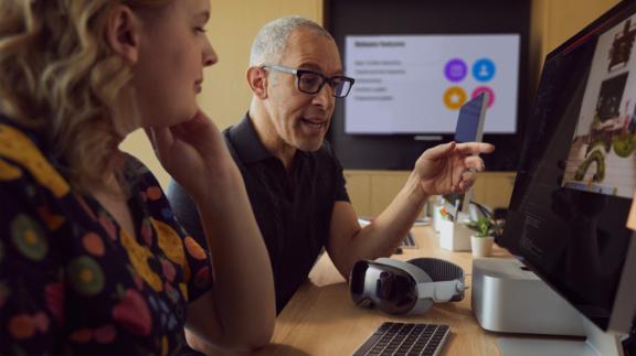 Vision desk b 2x Two coworkers review a desktop screen with a mixed reality headset on the desk