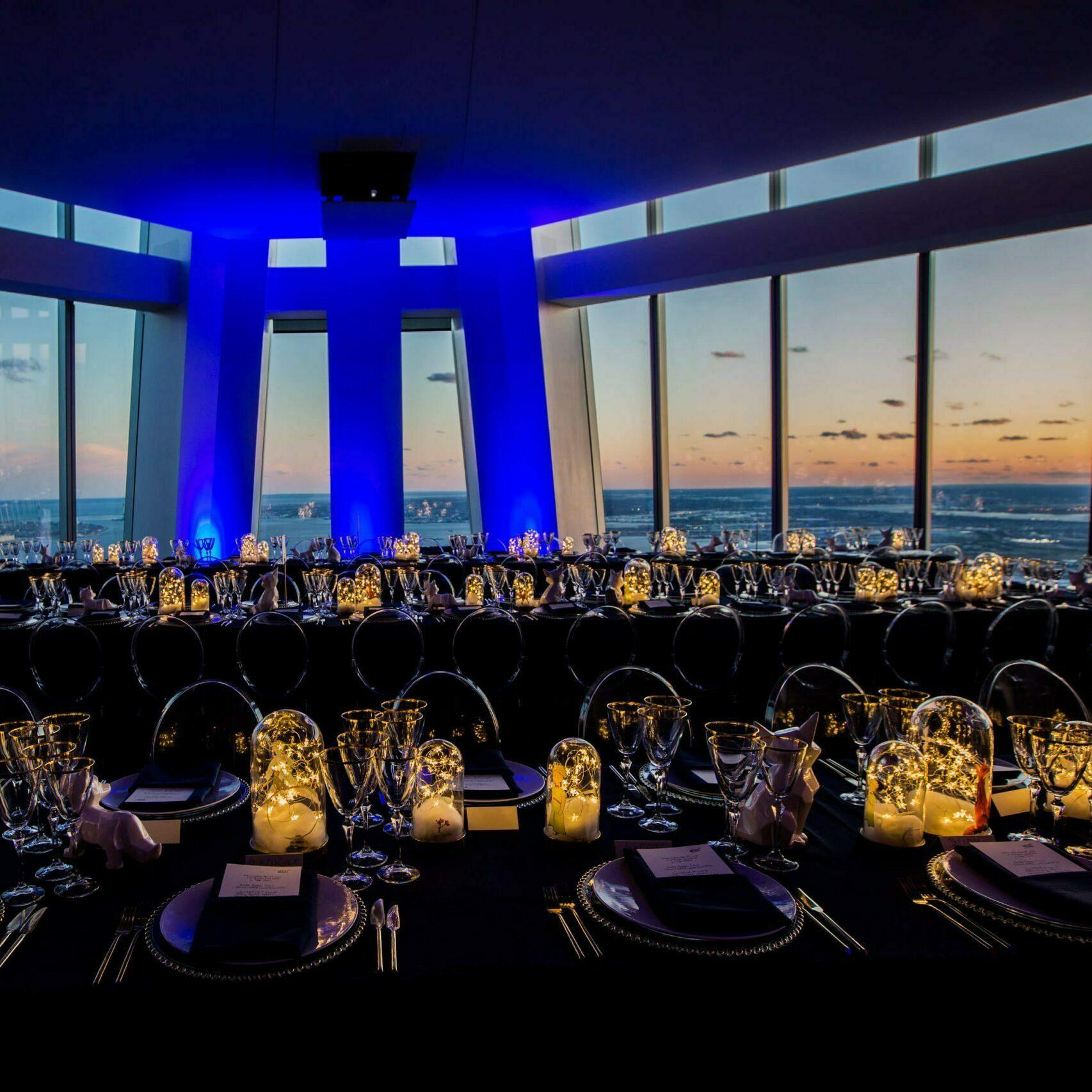 Gala dinner tables set in a high-rise event space overlooking the water at sunset