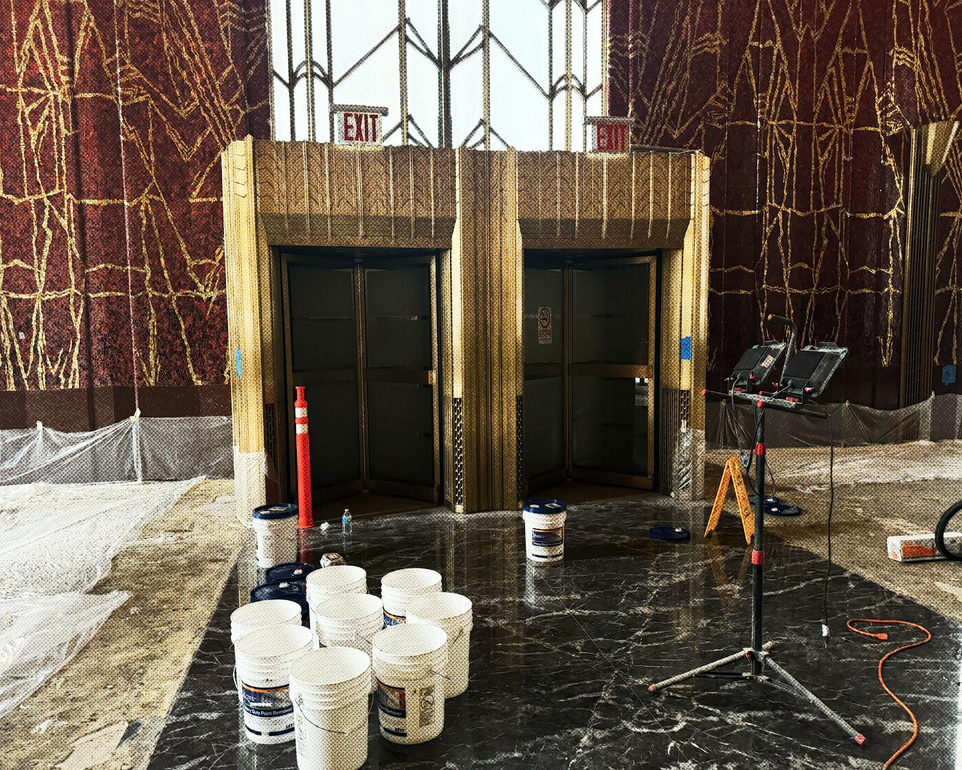 Elevator lobby under construction with paint buckets, work lights, and patterned walls.