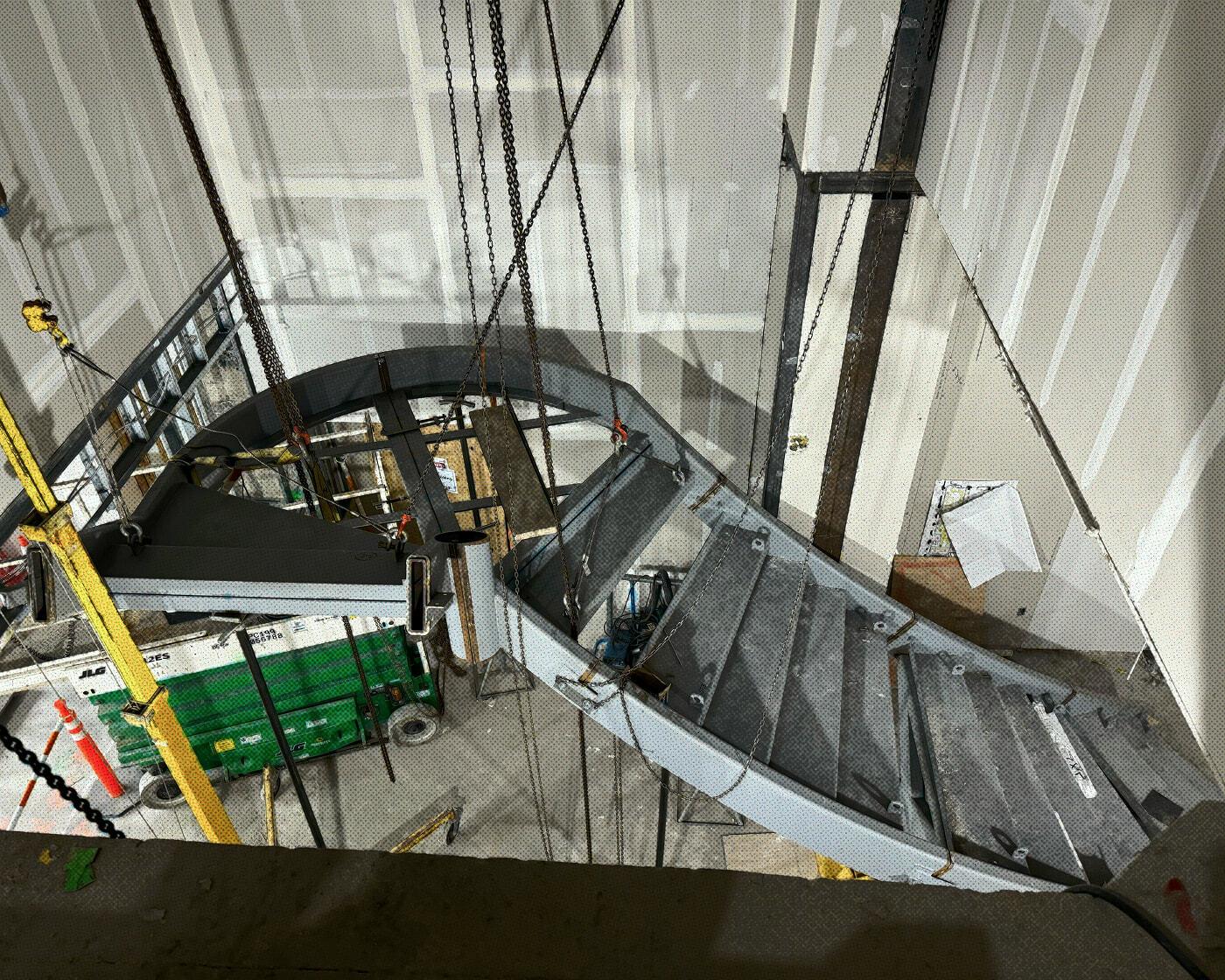 Curved staircase section hoisted on chains during installation in an unfinished interior.