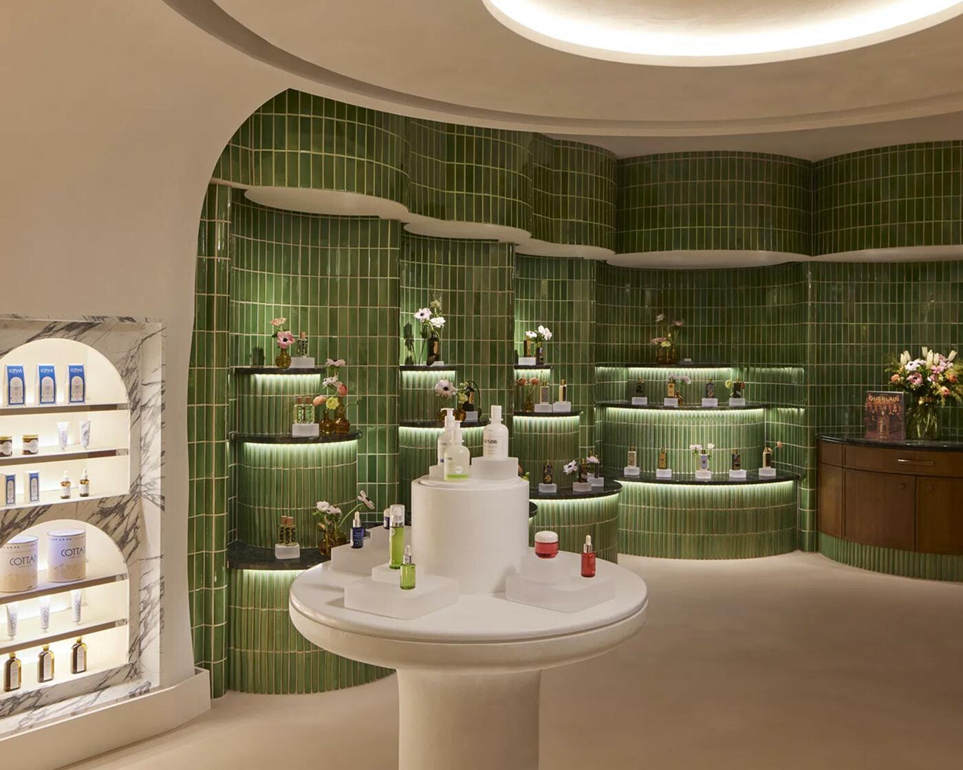 Green-tiled beauty product display with curved shelves, flowers, and a round white table.
