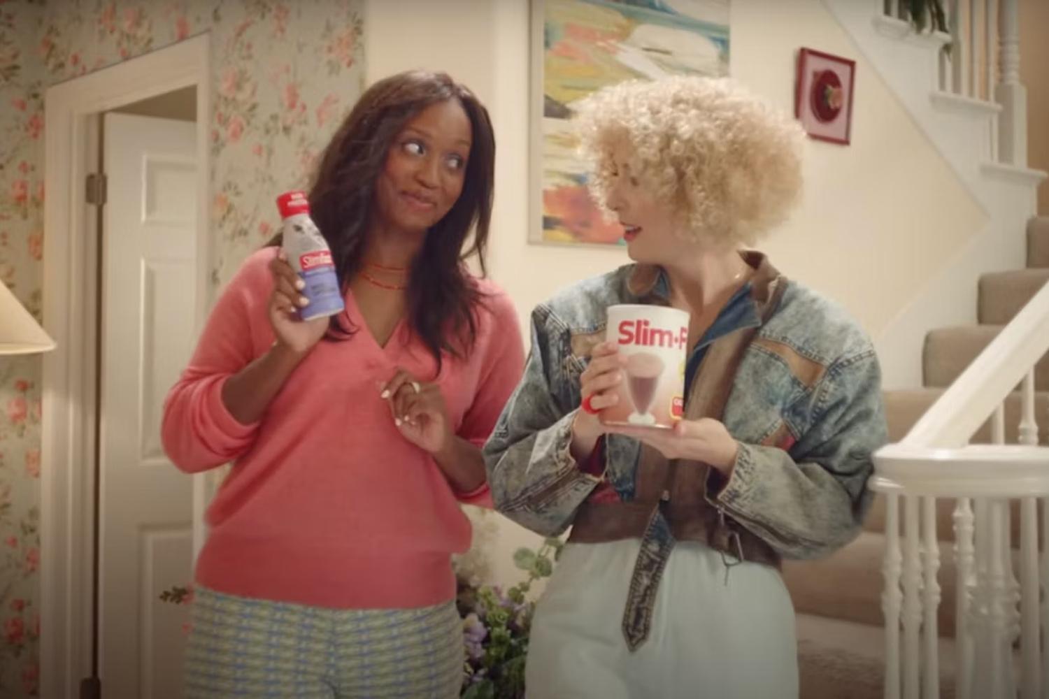 Two women compare meal replacement drinks in a home hallway near stairs