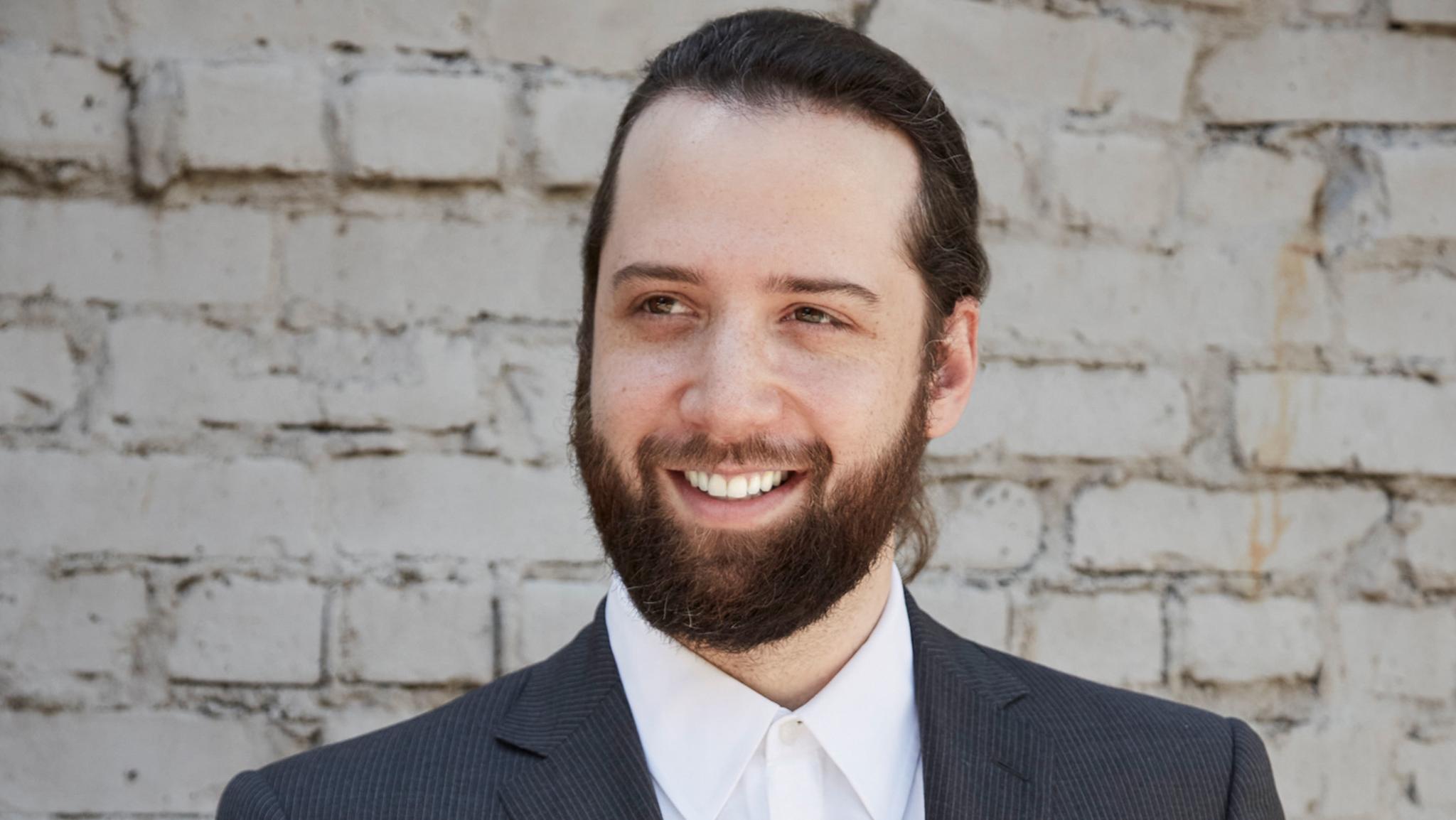 Smiling bearded man in a suit and white shirt against a white brick wall