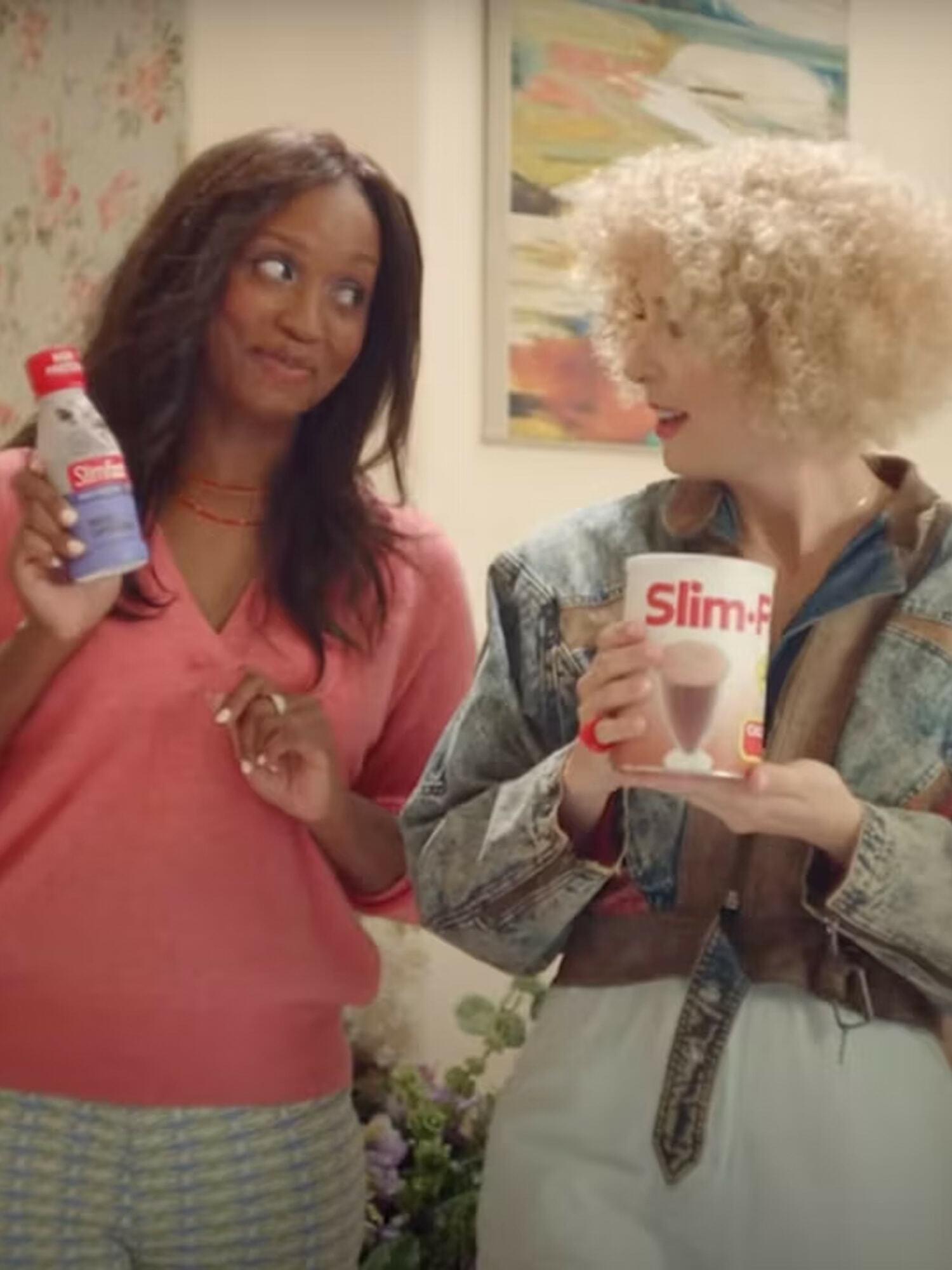 Two women holding SlimFast products while smiling at each other indoors