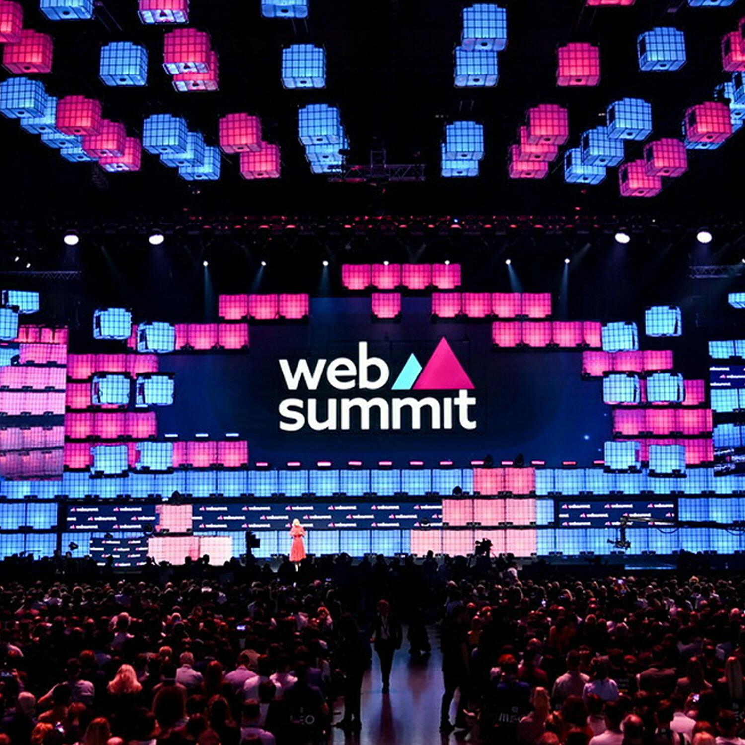 Web Summit main stage lit in pink and blue LED panels above a packed arena