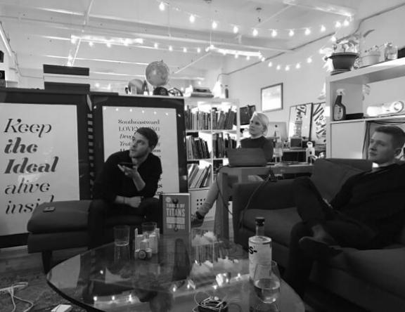 Screen Shot 2023 01 26 at 4 16 2 Black-and-white photo of three people in an office lounge under string lights, with a Keep the Ideal Alive Inside poster